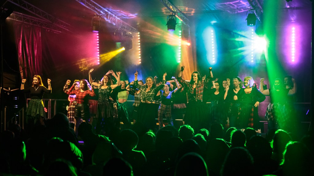 A colourfully lit stage during a musical theatre and dance performance with the heads of the audience highlighted by the stage light beams