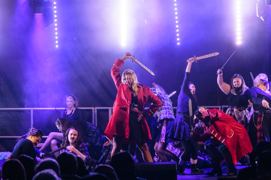 Bonnie Prince Charlie wields a sword on stage during an outdoor theatre performance at Dean Castle. The image is backlit with blue stage lighting and haze